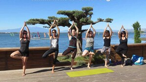 Un grupo de personas practicando yoga en una terraza de madera junto a un lago, con vista clara al agua y montañas distantes.