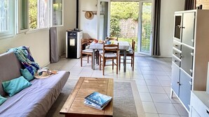 A spacious living area with a sofa, coffee table, and dining table. There is a kitchen area with cabinets and a refrigerator. The room has large windows and a view of greenery outside. - Peace, Beaches and Cycling on the Bay of Arcachon - Arès Cap Ferret  (Arès)