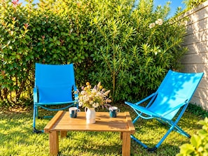 Two blue folding chairs, a wooden table, and a white pitcher with flowers.