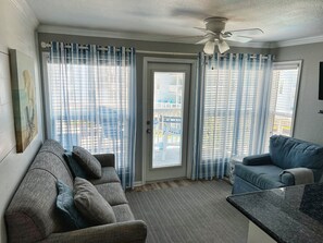 Ein Zimmer mit grauem Sofa, blauem Sofa, Deckenventilator und Blick auf Gebäude durch die Fenster.