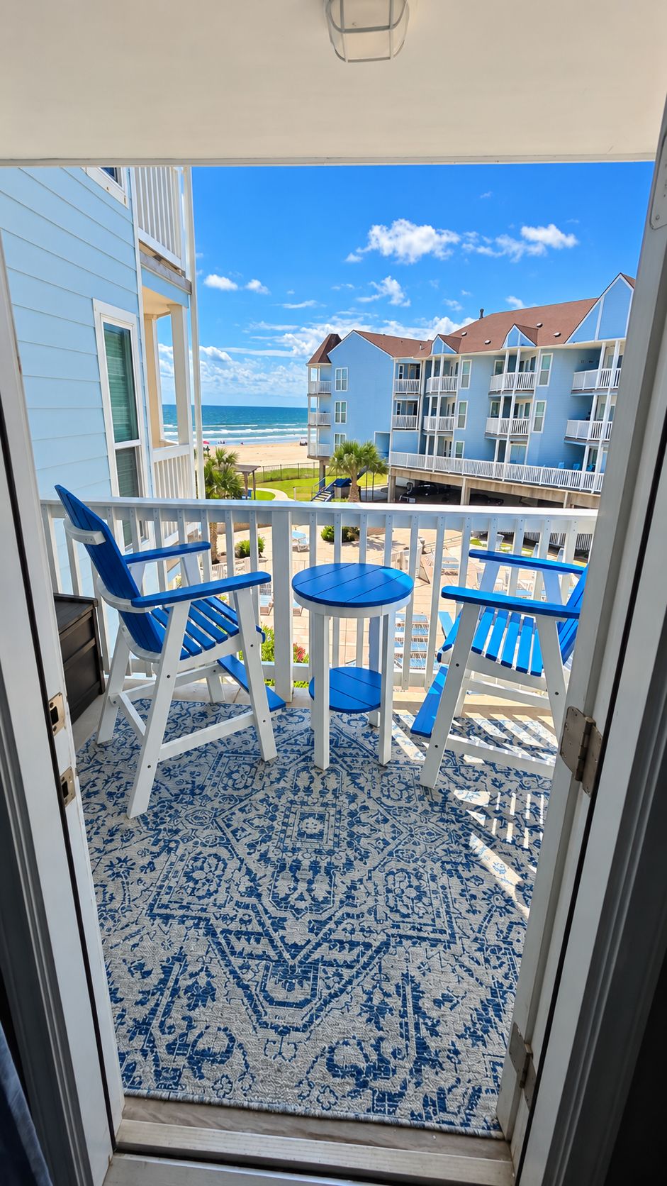 A balcony with blue chairs and a table, overlooking a beach and buildings.
