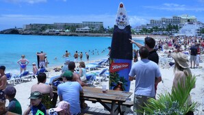 Une scène de plage avec une grande publicité pour une boisson, des chaises de plage et une table en bois.