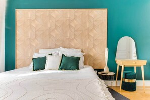 Una cama con ropa blanca y almohadas verdes, un cabecero de madera con un diseño geométrico, una mesita redonda con un florero y un aparador de madera con un espejo.