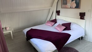 Ett sovrum med en vit säng, lila kuddar, en lila pläd och en tavla på väggen.