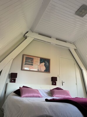 Un dormitorio con techo inclinado, una cama con ropa de cama blanca y rosa, dos altavoces fijados a la pared y un cuadro enmarcado sobre la puerta.