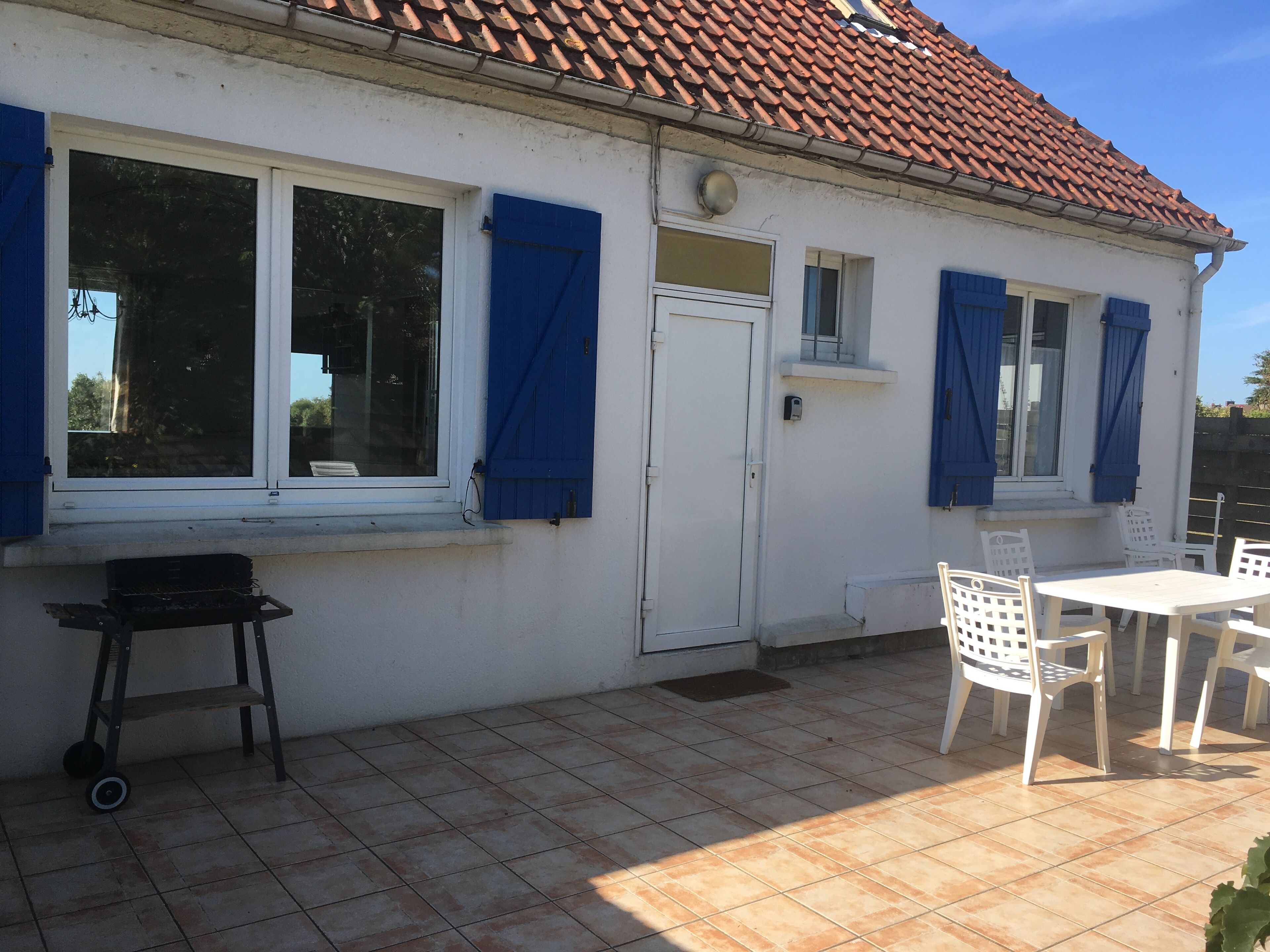 Een wit gebouw met een pannendak, grote ramen met blauwe luiken, een witte deur en een klein balkon met witte stoelen en een tafel.