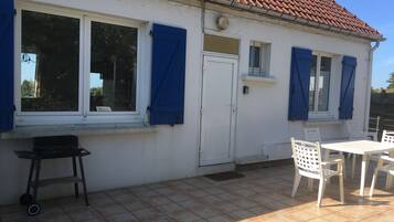 Een wit gebouw met een pannendak, grote ramen met blauwe luiken, een witte deur en een klein balkon met witte stoelen en een tafel.