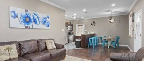 A living room with a brown leather sofa, a dining area with blue chairs, and a kitchen in the background.