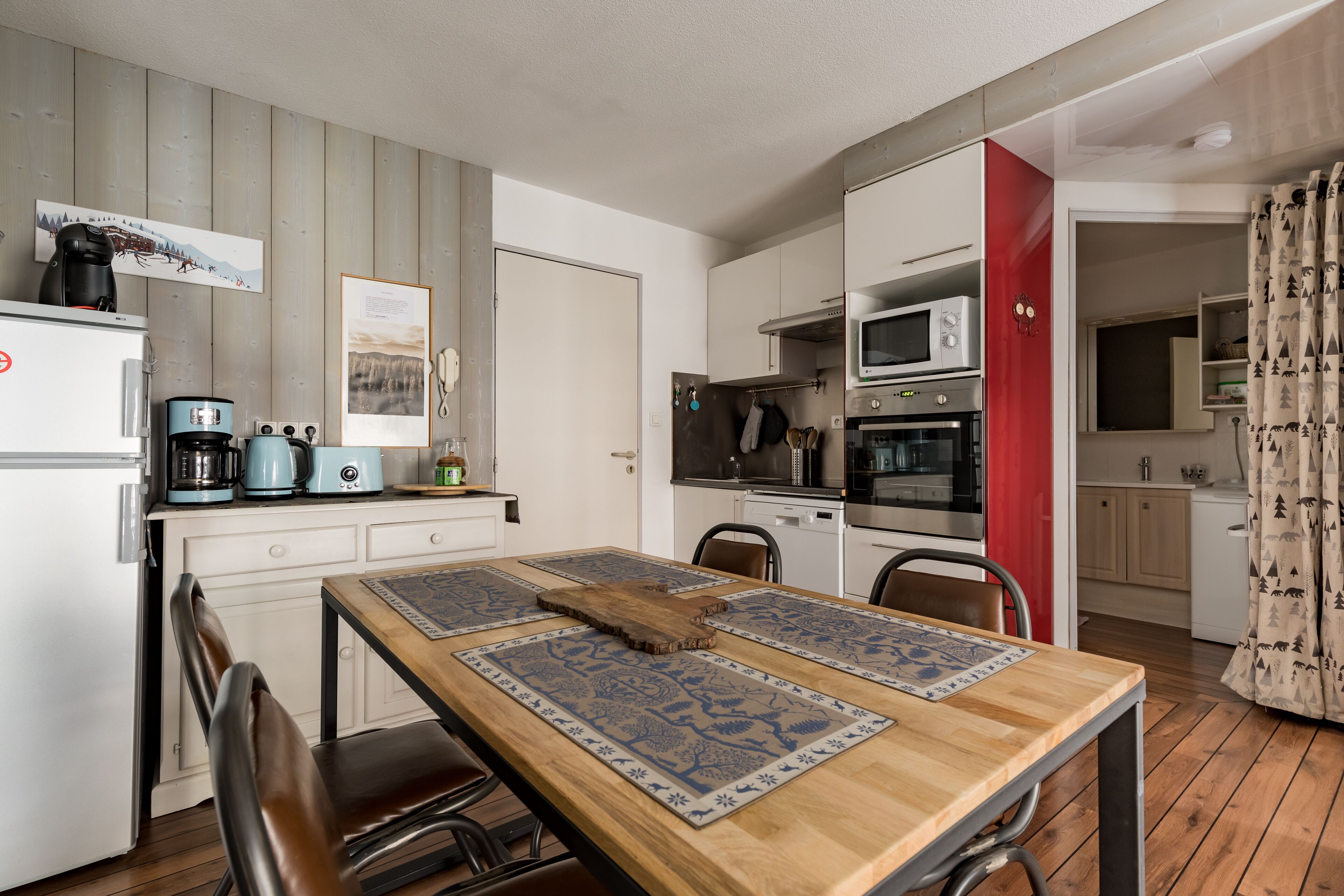 Een kleine keuken met witte kasten, een houten eettafel met blauwe placemats en een rode accentmuur.