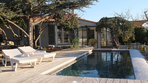 Eine Holzterrasse mit Liegestühlen, einem Schwimmbecken und einem Haus mit Ziegeldach.