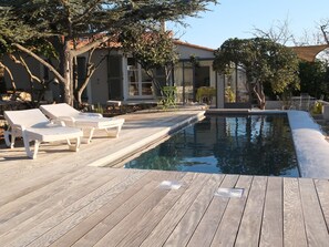 Una terraza de madera con sillones de descanso, una piscina y una casa con techo de tejas.