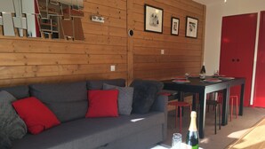 Una sala de estar moderna con un sofá gris, cojines rojos, una mesa de centro blanca con una botella y vasos, y una pared de madera con marcos.