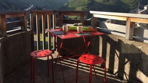 Una mesa metálica roja con dos taburetes, una taza blanca y una barra de pan en un balcón con vistas a un paisaje montañoso.