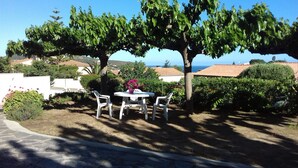 A patio with a round table and four chairs, surrounded by lush greenery and a view of the sea. - Ground floor villa flat in Monticello (Monticello)