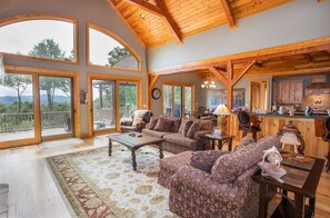 Relax in a spacious living area with a high ceiling and exposed beams, featuring a curved glass door leading to an outdoor deck with scenic views.