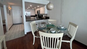 Uma área de refeições com uma mesa de vidro posta para quatro pessoas, rodeada por cadeiras brancas. Um candeeiro pendente é visível acima da mesa.