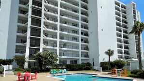 Uma piscina com escada de metal, rodeada por um edifício branco com várias varandas e uma palmeira.