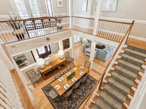 A spacious loft-style apartment with a high ceiling, a dining area with a wooden table and chairs, a living room with a sofa and a fireplace, and a staircase with a railing.