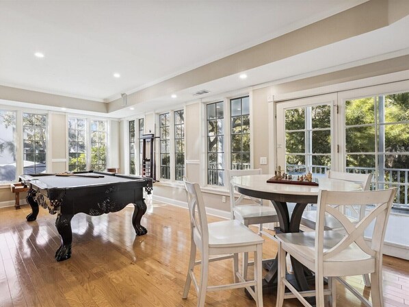 A spacious room with a pool table, a dining table with chairs, large windows, and a view of greenery outside.