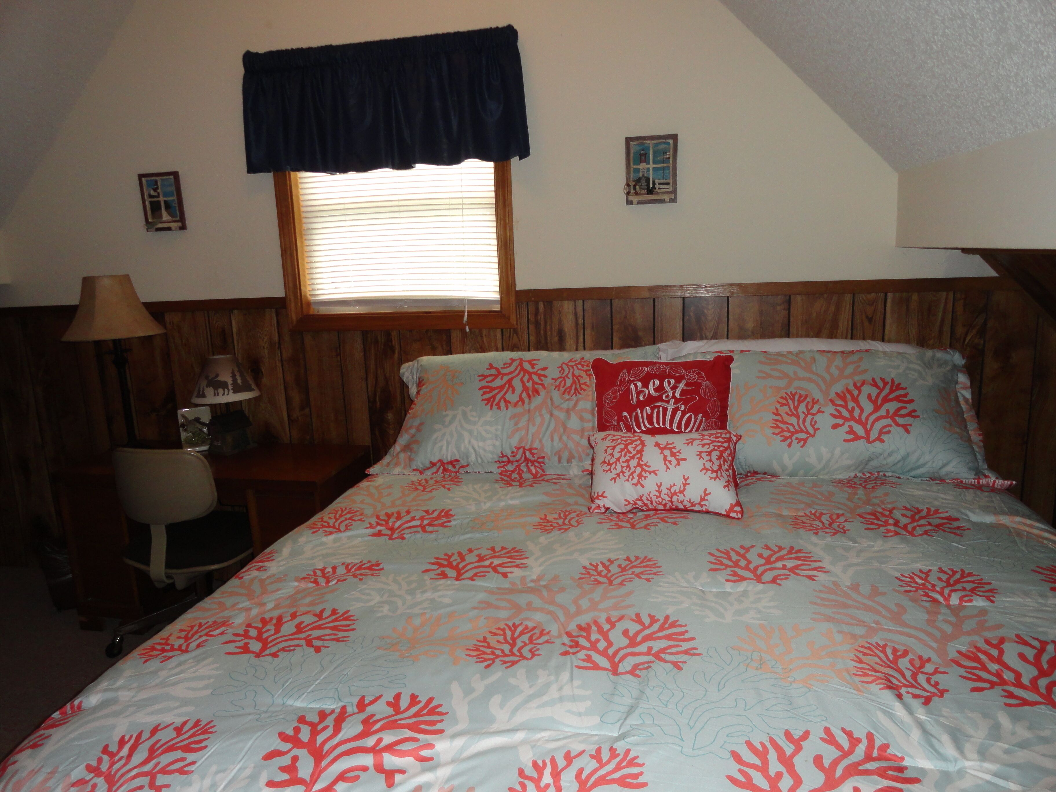 A bedroom with a bed, a nightstand, a lamp, a window with blinds, and a small wall decoration.