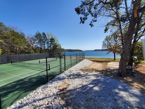 En tennisbane med net, omgivet af grus og træer, med udsigt over vand.