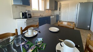 A modern kitchen with blue cabinets, a stainless steel refrigerator, and a dining table set with plates, cups, and a coffee maker. - Farniente & sunsets on the Gulf of Ajaccio and the Sanguinaires Islands (Bastelicaccia)
