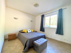 A bedroom with a bed, a bedside table, a bench, a window with curtains, and a ceiling light.