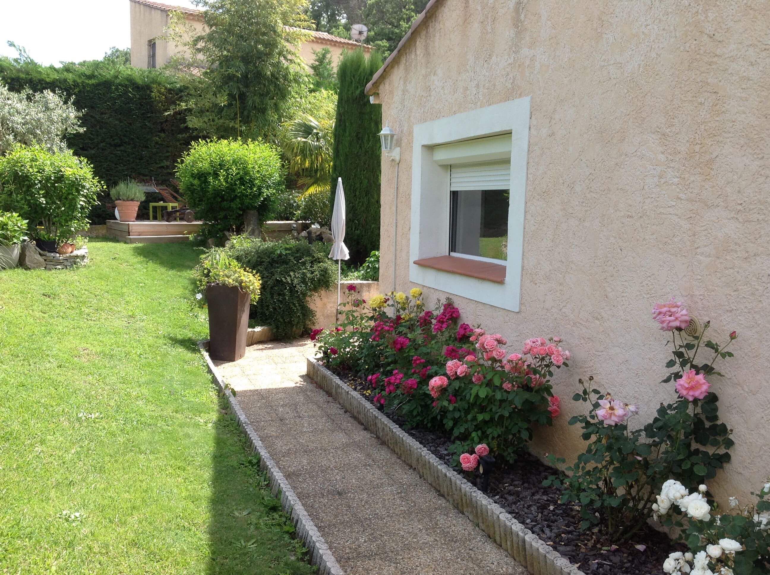 Ein Garten mit einem Weg, der zu einem Haus mit einem Fenster führt. Um das Haus herum befinden sich verschiedene Pflanzen und Blumen.