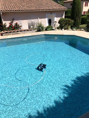 En klar, blå swimmingpool med en flisekant, omgivet af en stenvæg og en bygning med et tag dækket af fliser.