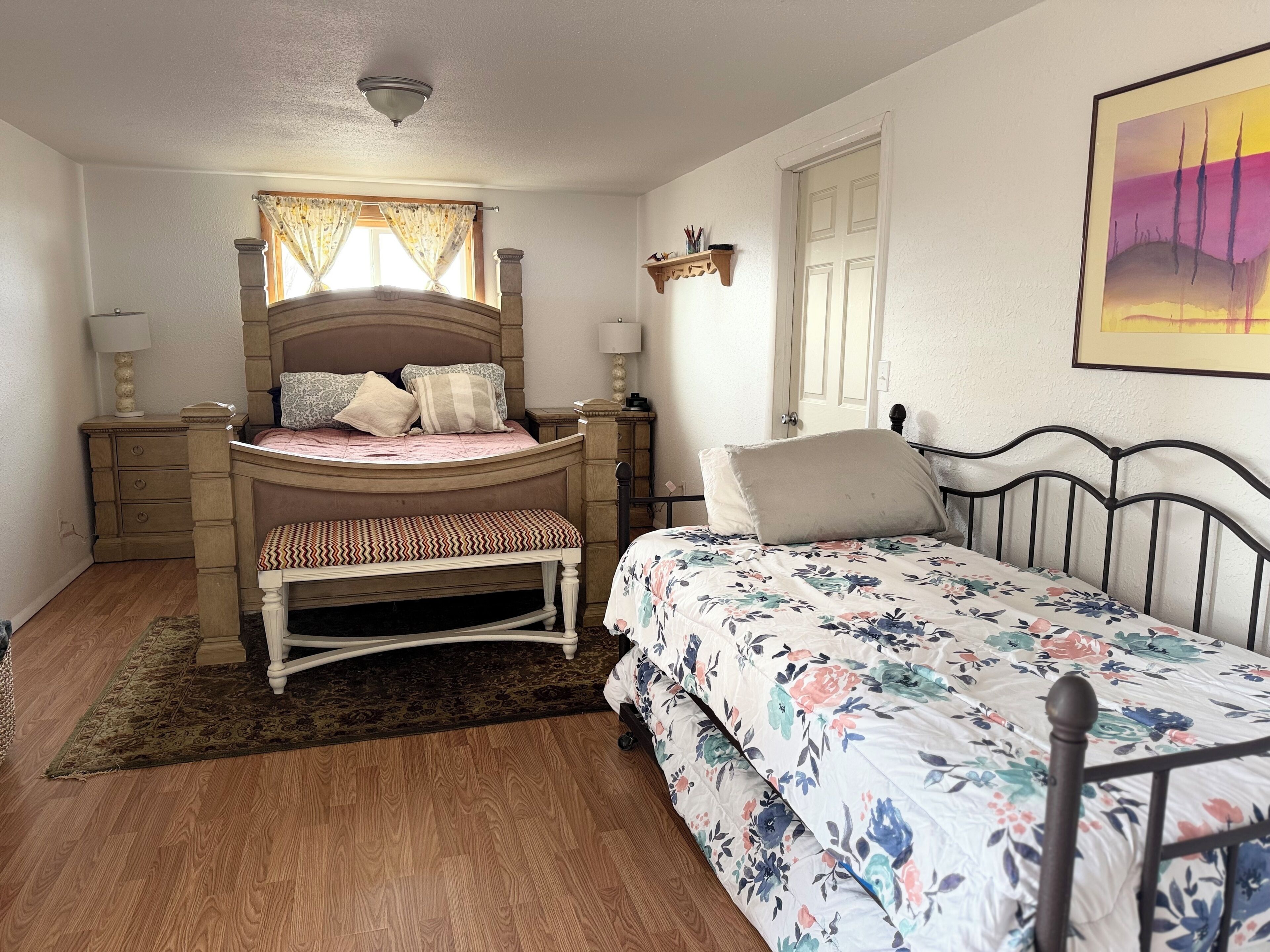 A bedroom with a wooden bed, a bench, a nightstand, a lamp, a window with curtains, and a door.