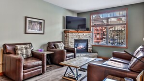 A living room with leather sofas, a stone fireplace, a flat-screen TV, and a large window with a view of buildings. - Inviting Condo – Hot Tub & Relaxing Atmosphere! (Canmore)