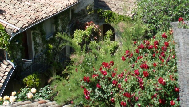 Um pátio com edifícios de pedra, uma torre de sino e flores vermelhas.