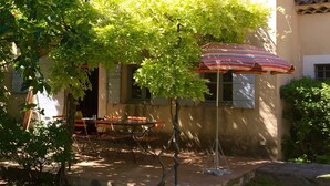 Eine Terrasss mit Tisch, Stühlen, einem gestreiften Sonnenschirm und einem Baum, der Schatten spendet.