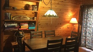 Een gezellige eethoek met een houten tafel en stoelen, een hanglamp en een boekenkast vol boeken en decoratieve spullen.