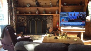 Een gezellig chaletinterieur met een open haard, houten planken met decoratieve objecten, een televisie waarop nieuws wordt vertoond, en een comfortabele zithoek.