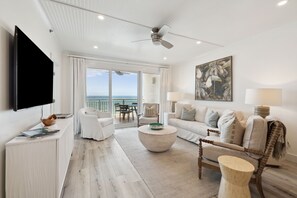 A spacious living room with a large flat-screen TV, a comfortable sofa, a coffee table, and a view of the ocean through large windows.