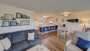 A modern living room with a grey sofa, a coffee table, and a dining area with a white table and chairs. A kitchen with a blue island and white cabinets is visible in the background. - Beach Please- Oceanfront easy access to the beach (Long Beach)