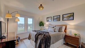 A bedroom with a large bed, bedside lamps, a dresser, a window, and framed pictures on the wall. - Beach Please- Oceanfront easy access to the beach (Long Beach)