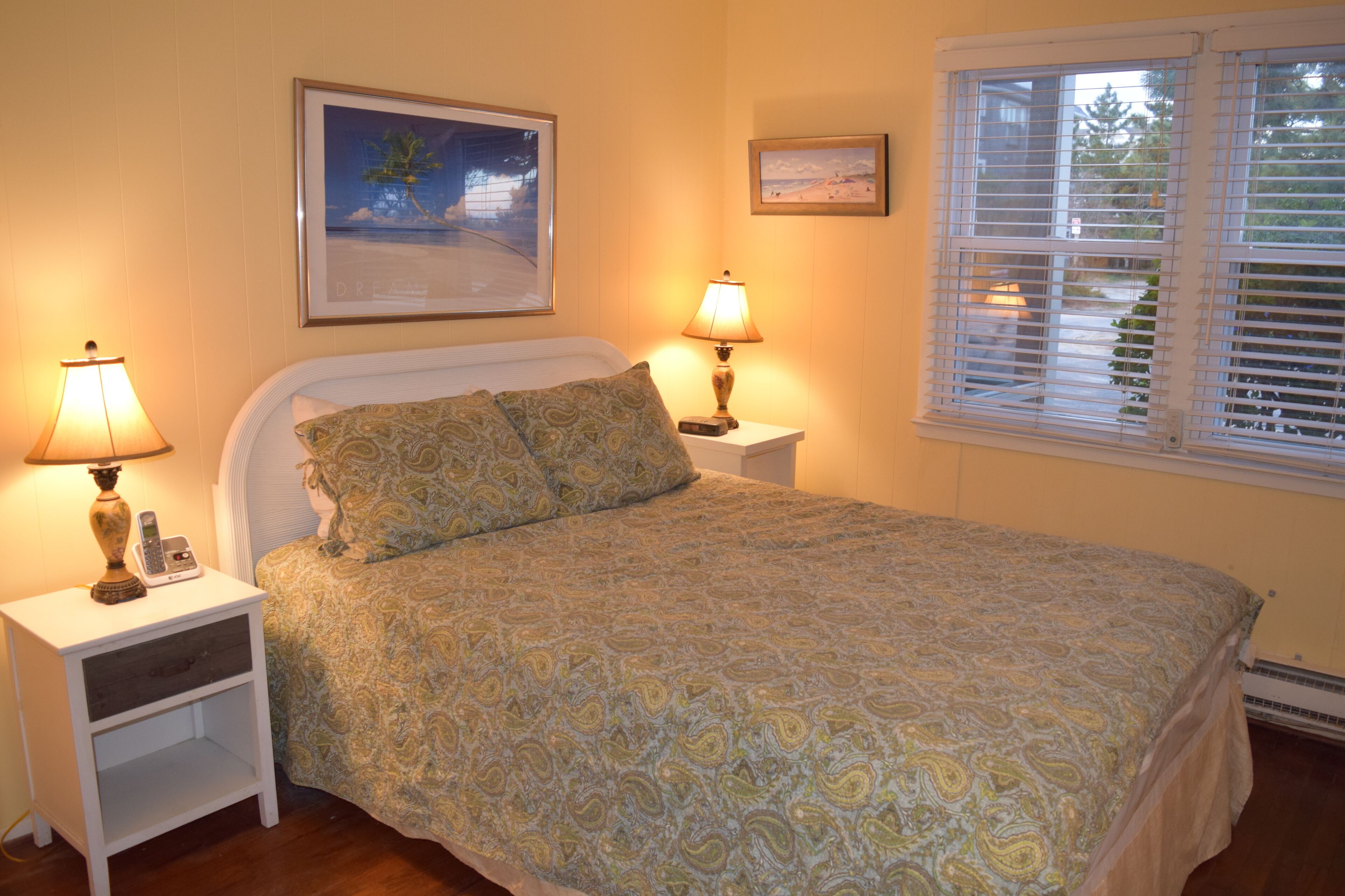Une chambre à coucher comprenant un lit, deux tables de chevet, une commode et une fenêtre avec des stores.