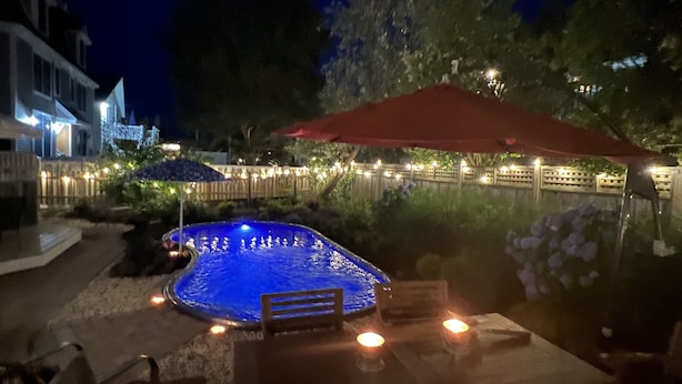 Piscina iluminada com luz azul, rodeada por um deck de madeira, um guarda-sol vermelho e uma cerca com luzes de cordão.