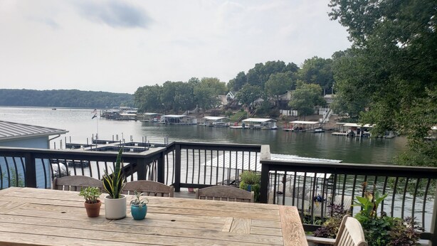 Une terrasse en bois avec une table et des chaises, donnant sur un lac où l’on aperçoit des bateaux et des maisons au loin.