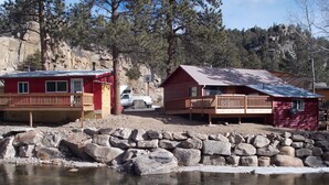 Une cabane dotée d’une terrasse en bois et d’un petit porche, située à proximité d’une côte rocheuse bordée d’une étendue d’eau.