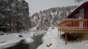 Une cabane dotée d’une terrasse en bois offrant une vue sur un paysage enneigé et un ruisseau.