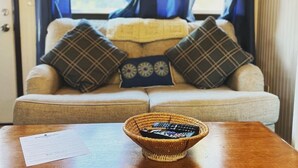 Un salon chaleureux avec un canapé agrémenté de coussins à carreaux, une table en bois sur laquelle se trouve un panier contenant des télécommandes, et une fenêtre ornée de rideaux bleus.