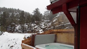 Une terrasse en bois avec un jacuzzi, entourée d’un paysage enneigé et d’une colline rocheuse parsemée d’arbres.