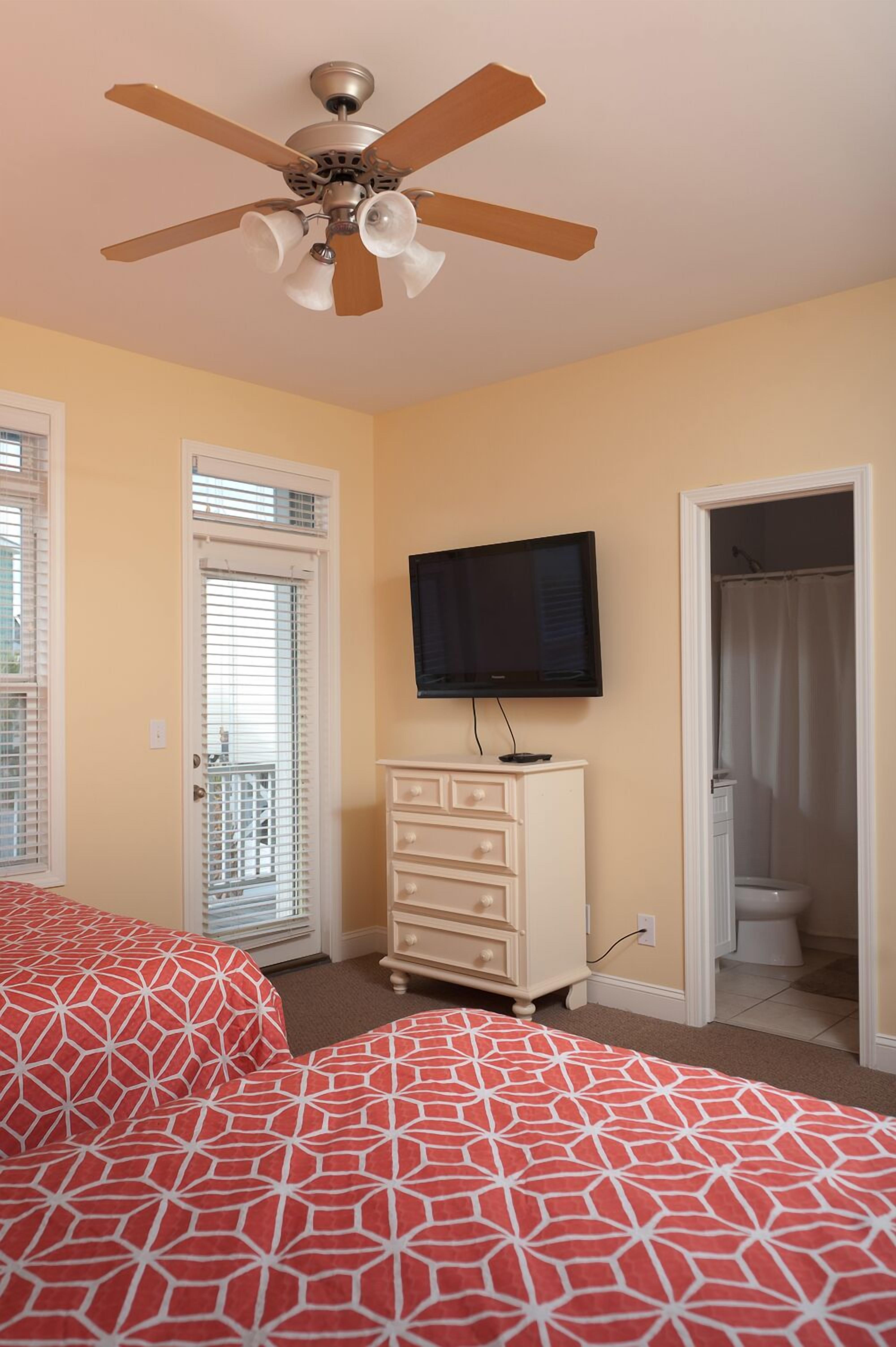 A bedroom with a bed, a ceiling fan, a television, a dresser, and a bathroom door.