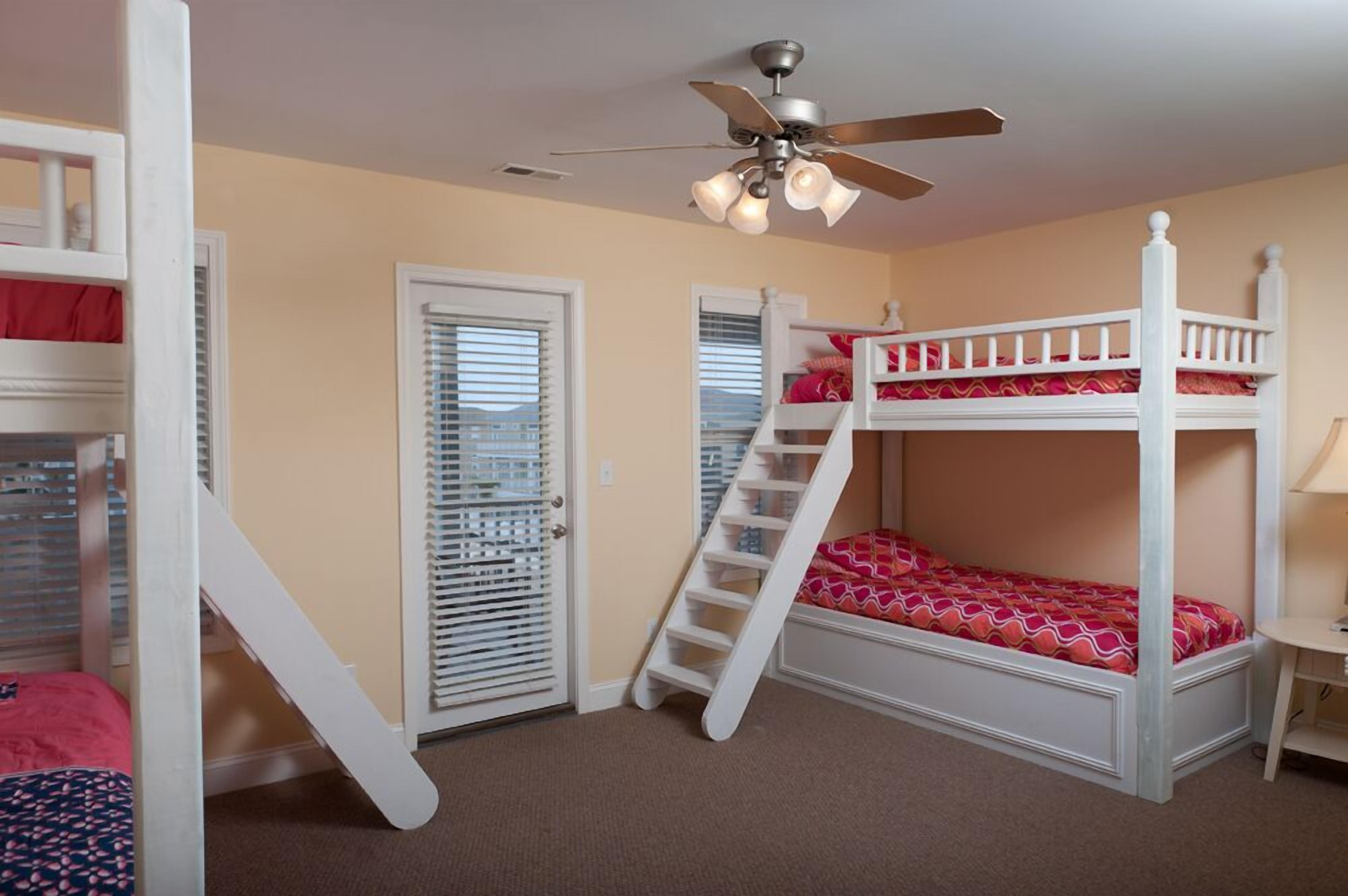 A bunk bed room with a ceiling fan, a door with blinds, and a small table.