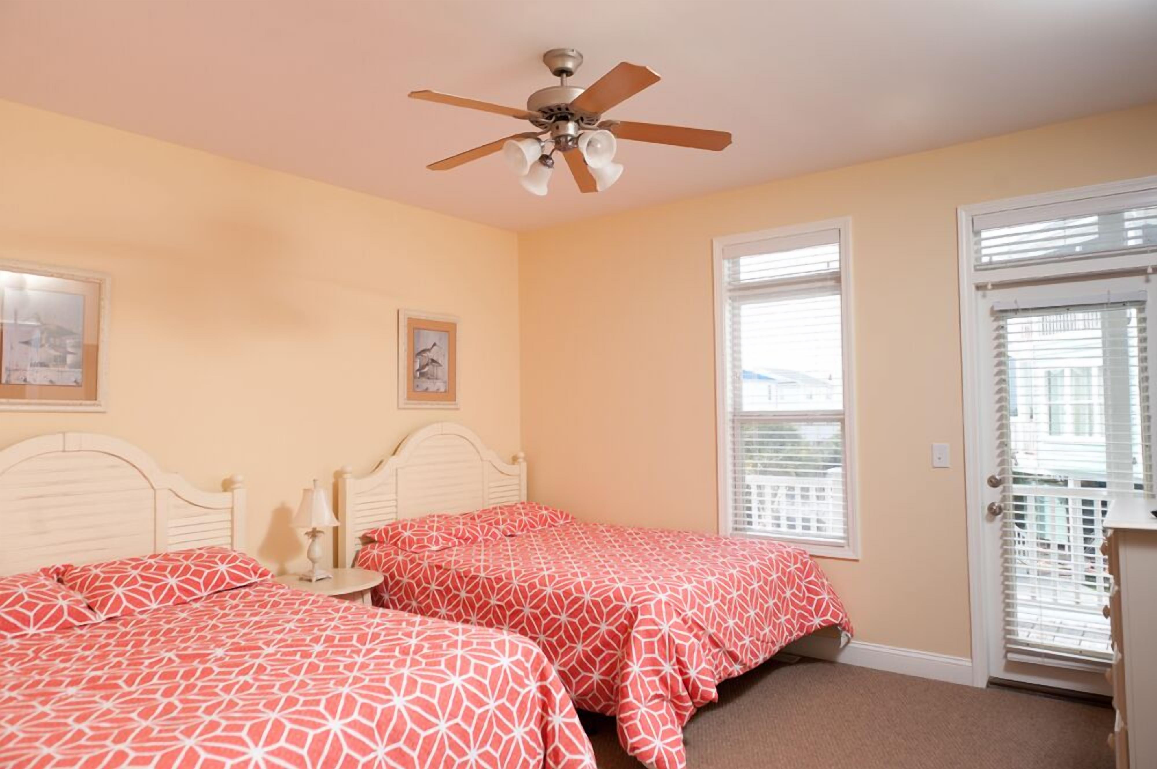 A bedroom with two beds, a ceiling fan, a window with blinds, and a door with a view of the outside.