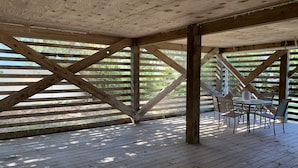 A covered outdoor area with a wooden deck, a table with chairs, and a view of a green landscape through wooden slats.