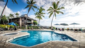 Ein Pool mit Liegestühlen und Sonnenschirmen, umgeben von Palmen und einem Gebäude mit Balkon.